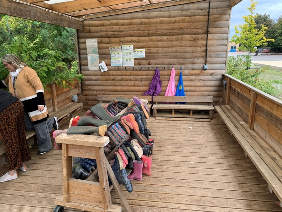 An outdoor wooden shelter with benches, hooks for hanging items, and stacks of wellington boots, with two adults near a wall display.